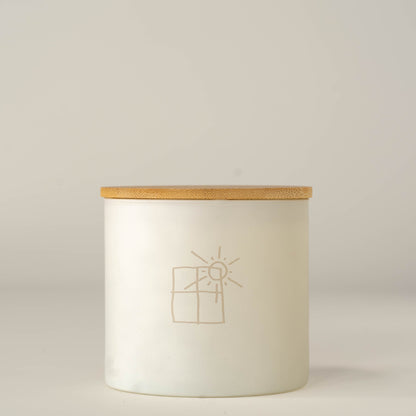 A white glass jar candle with a light natural wood lid, featuring a minimalist beige line drawing of a sun and window on the front, set against a neutral light gray background.